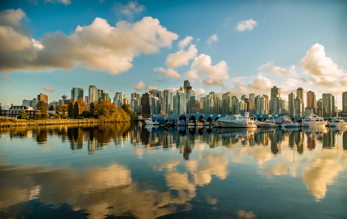 Scenic Vancouver skyline reflecting on calm water, showcasing modern architecture, docked boats, and a partly cloudy sky at sunset.